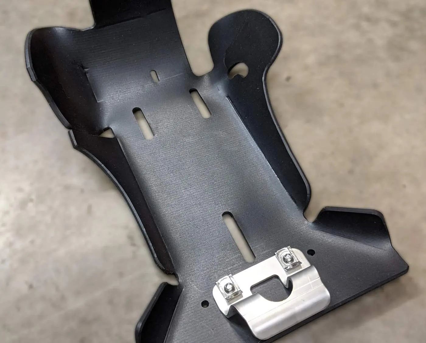 Black plastic UHMW dirt bike skid plate with metal fastener on a beige surface