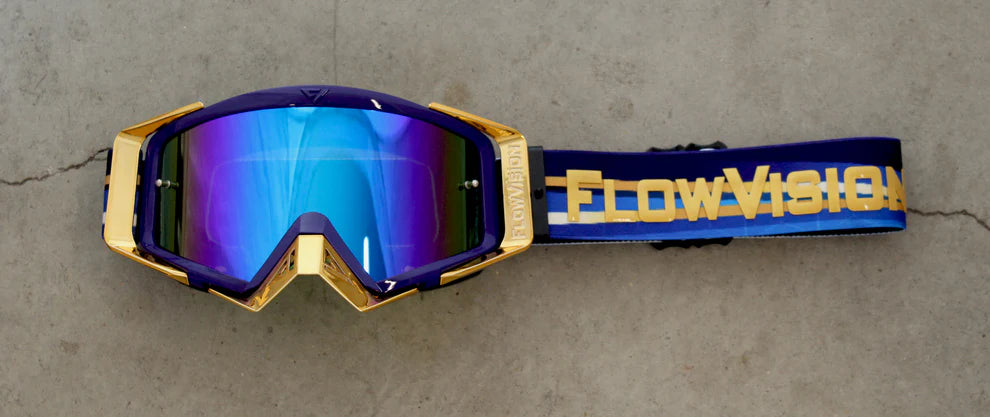 Gold goggles with blue lenses and 'FlowVision' strap on a gray surface