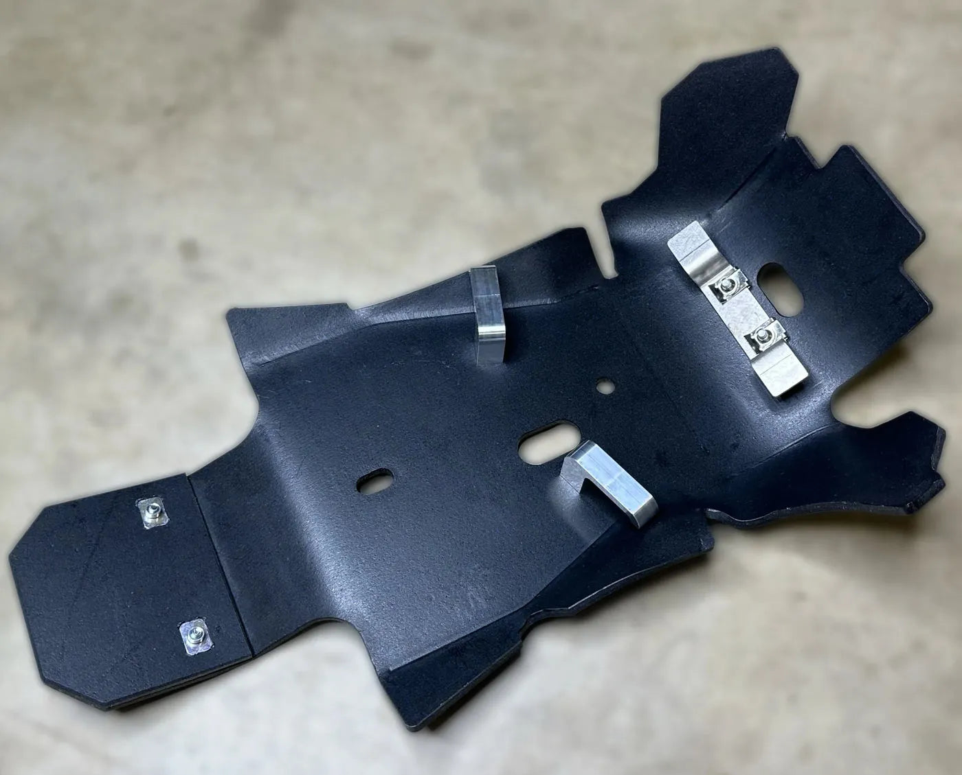 Black UHMW plastic dirt bike skid plate and metal bracket on a beige surface