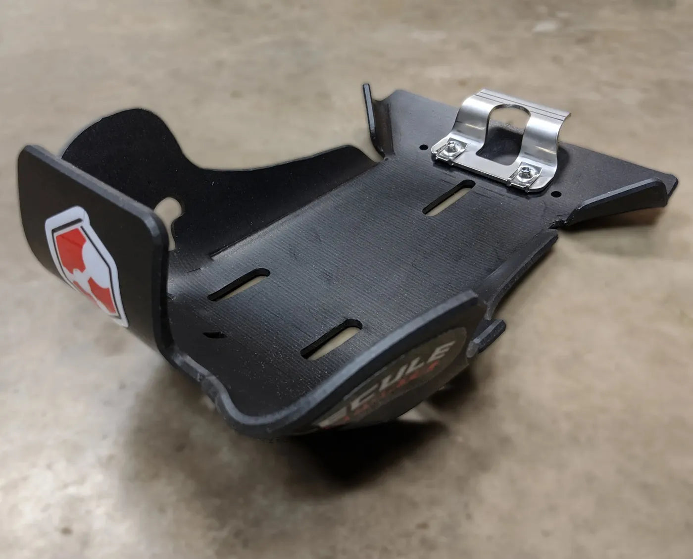 Black dirt bike skid plate with a metal bracket and logo on a beige surface