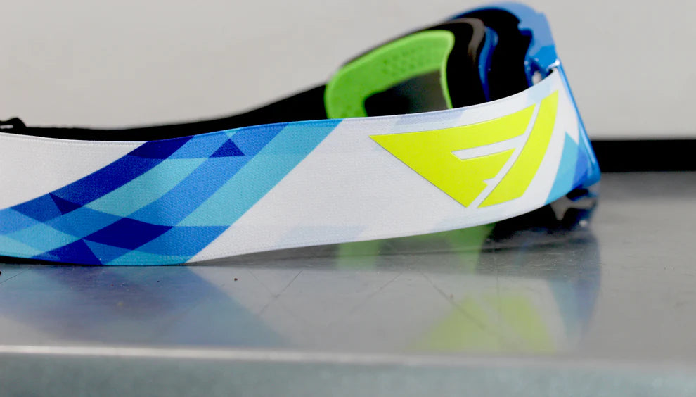 Close-up of a colorful MX goggle with blue, green, and white design on a reflective surface.
