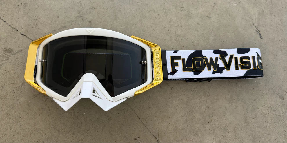 White and gold goggles with a camouflage strap on a concrete surface, featuring 'FlowVisor' branding.