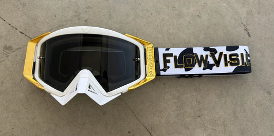 White and gold goggles with a camouflage strap on a concrete surface, featuring 'FlowVisor' branding.