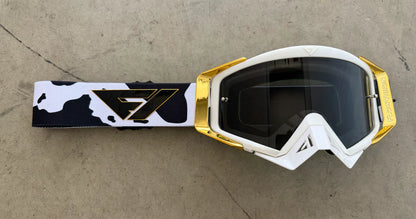 White goggles with gold accents and a black and white patterned strap on a concrete surface