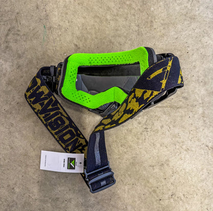 Black goggles with green face foam and a black and gold strap on a concrete surface