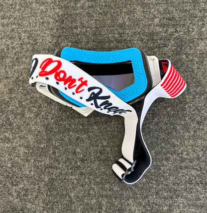 Blue and white motocross goggles with 'I Don't Kneel' strap on a gray background
